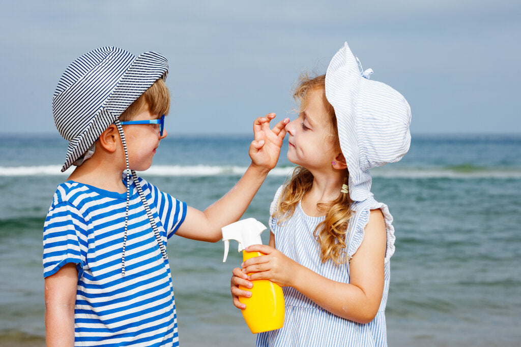 Two children at the beach applying sunscreen and wearing sun hats, showing a playful approach to building a sun protection wardrobe.
