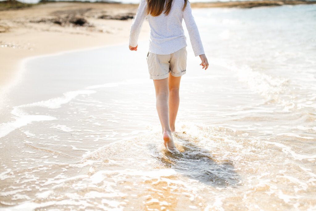 Child in a loose white long-sleeve shirt and shorts walking barefoot through the surf on a bright, sunny beach, covered up and cool in direct sun.