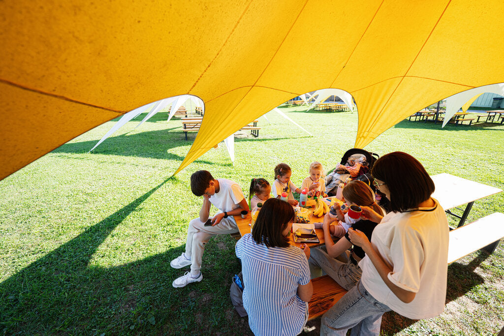 Family sitting under a large shade canopy at a park picnic table on a sunny day, illustrating how shade can reduce direct sun exposure but may not block all reflected UV rays.