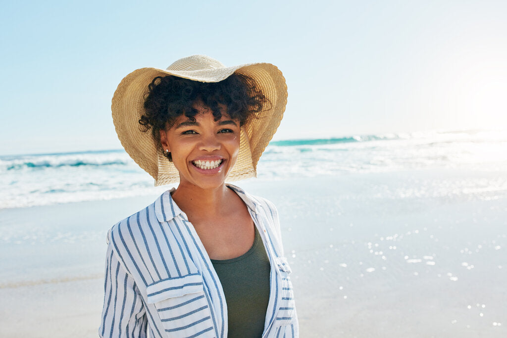 Person standing by the ocean on a clear day, wearing a sun hat and light layers, highlighting that UV exposure and sunburn risk can occur year-round, not just during summer.