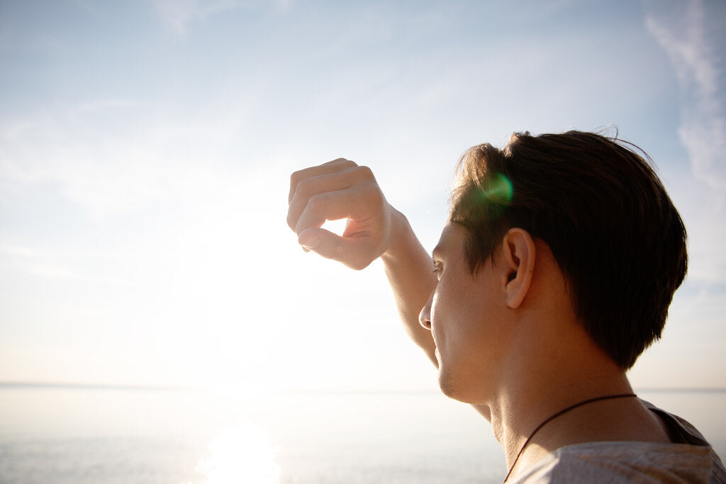 Man shielding his eyes from the bright sun near the water, illustrating the need for UVB protection and daily sun safety habits.