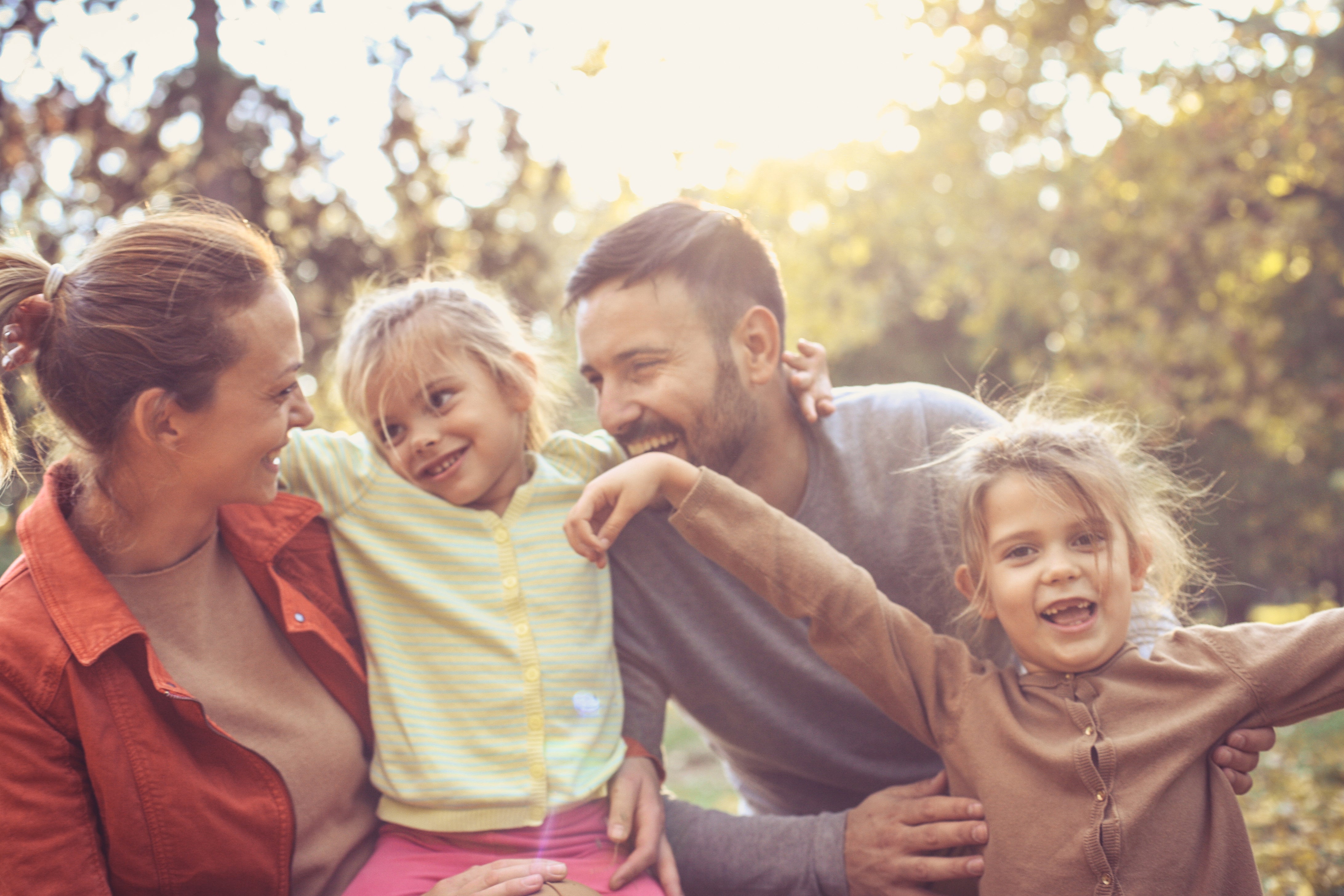 A happy family of four enjoying a sunny fall day outdoors. The parents smile as their two young daughters laugh and play in front of them, surrounded by soft sunlight and trees.