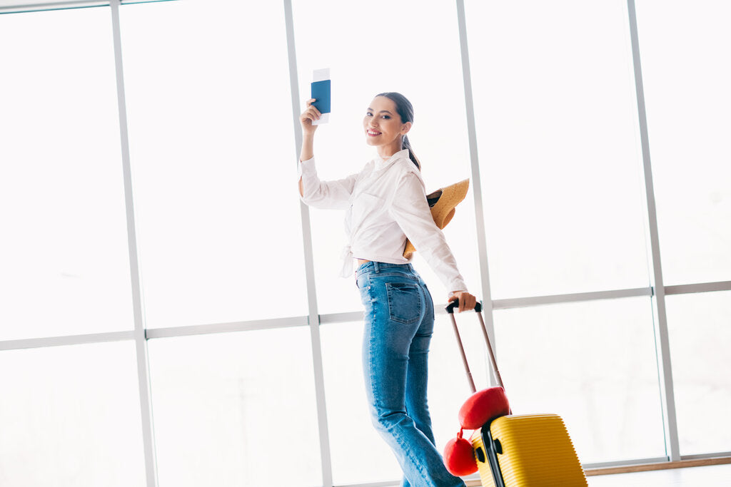 Smiling woman walking through an airport with a suitcase and boarding pass, ready for a trip to a high UV travel destination, emphasizing the importance of packing a sun protection wardrobe.