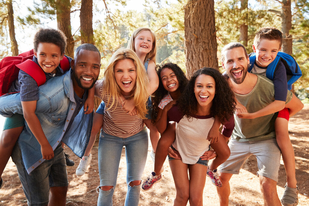 A happy, diverse group of families enjoying an outdoor adventure. Two adults give piggyback rides to children, all smiling and laughing, surrounded by trees on a sunny day.
