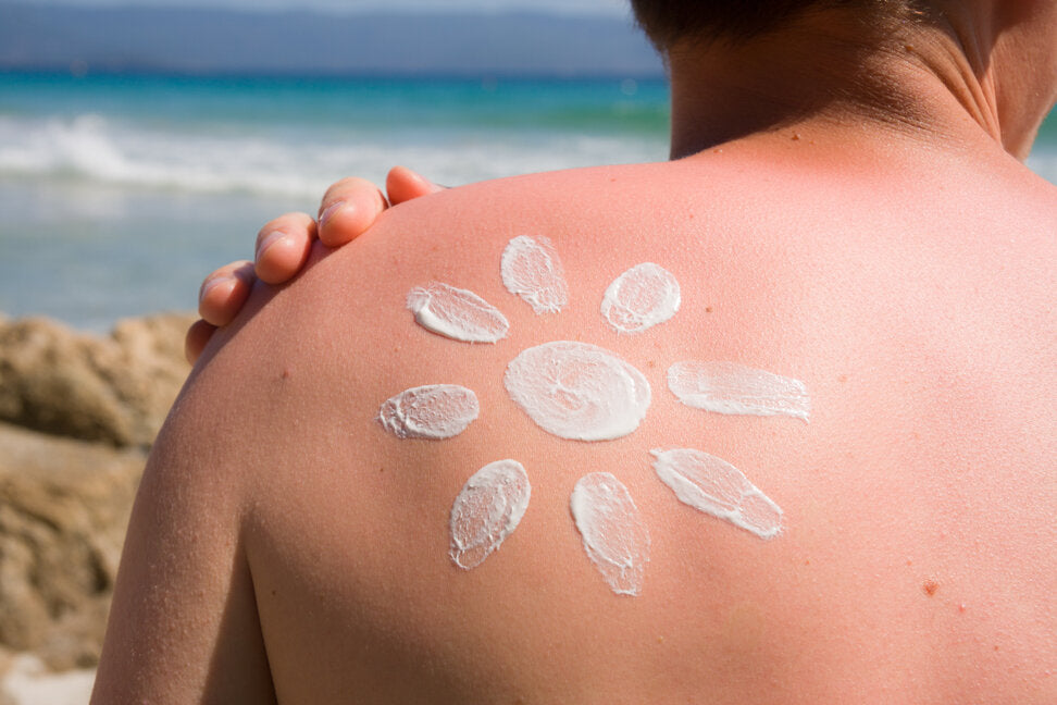 Close-up of a person’s sun-exposed back with sunscreen applied in the shape of a sun, standing near a beach with the ocean in the background.