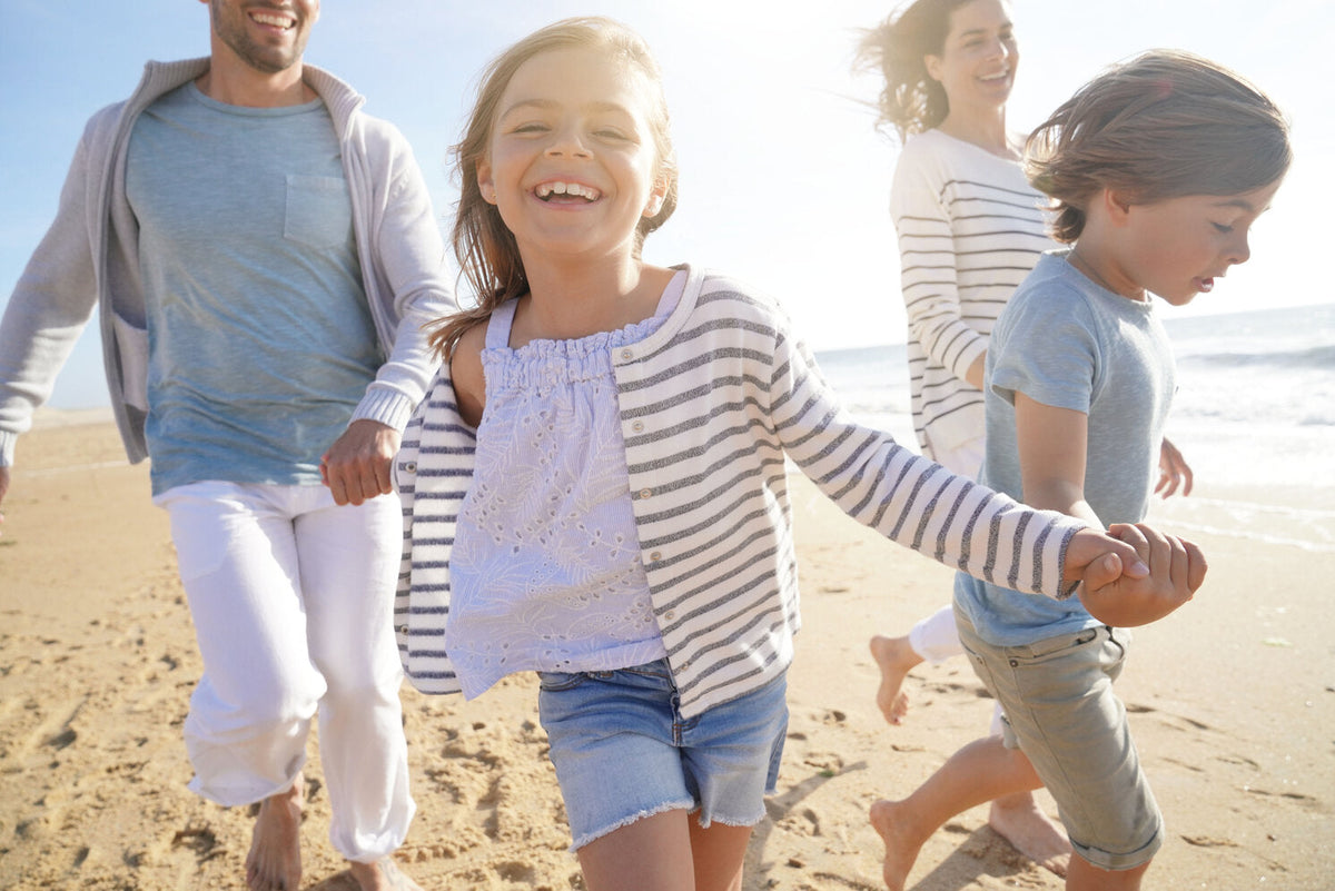 A joyful family runs on the beach, with the children and parents wearing light clothing, enjoying the sunny day safely under the sun's rays.