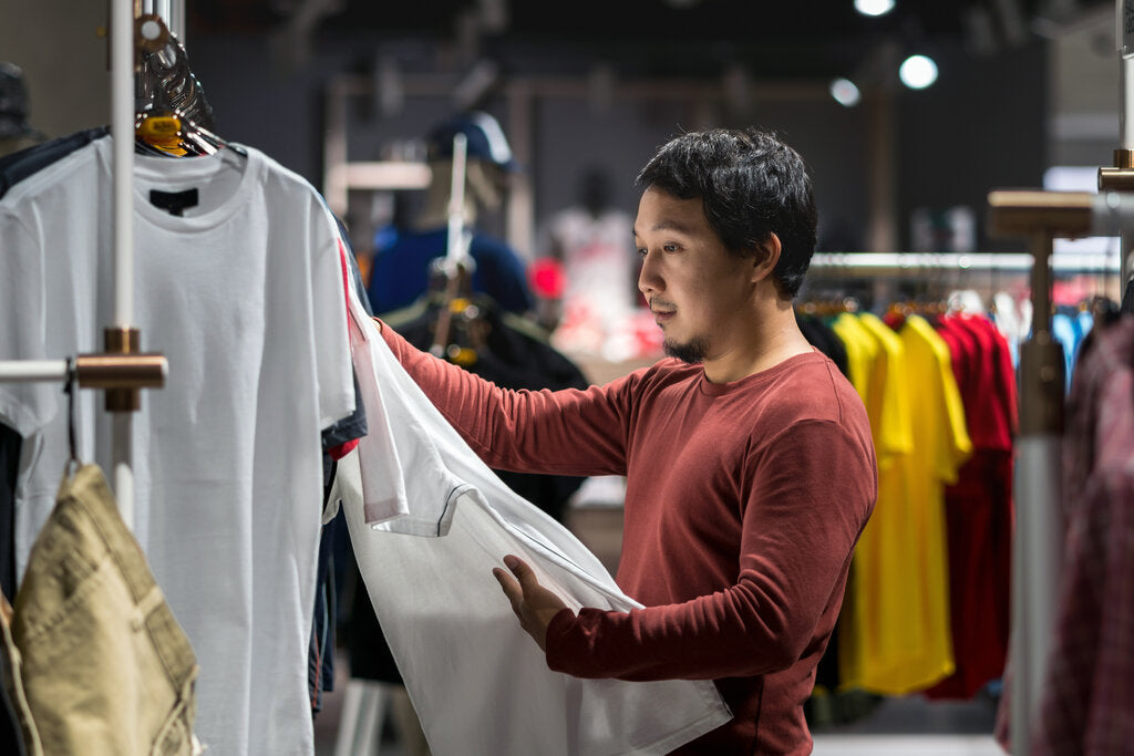 Man examining a white t-shirt in a clothing store, illustrating the importance of checking labels to identify UV protection and UPF-rated clothing.