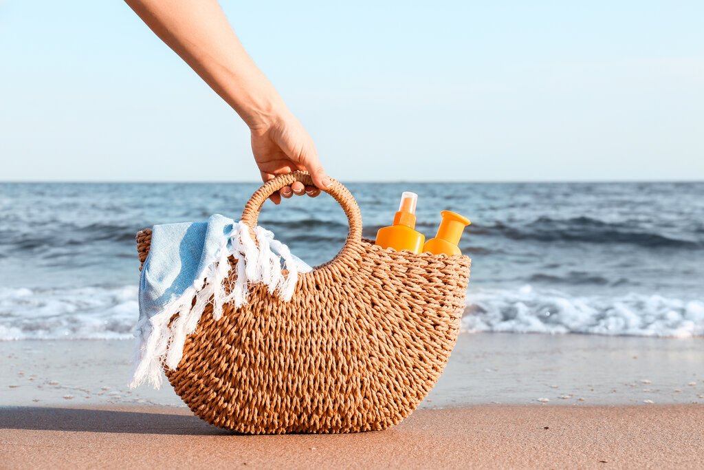 Wicker beach bag with sunscreen bottles and a towel, placed on the sand near the ocean—reminding us to pack sun protection essentials for a safe day outdoors.