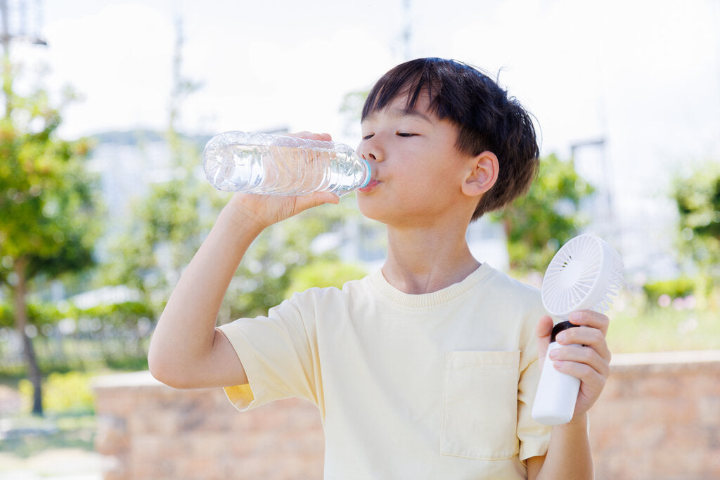 Young boy staying hydrated during a heat wave, drinking water and holding a portable fan outdoors