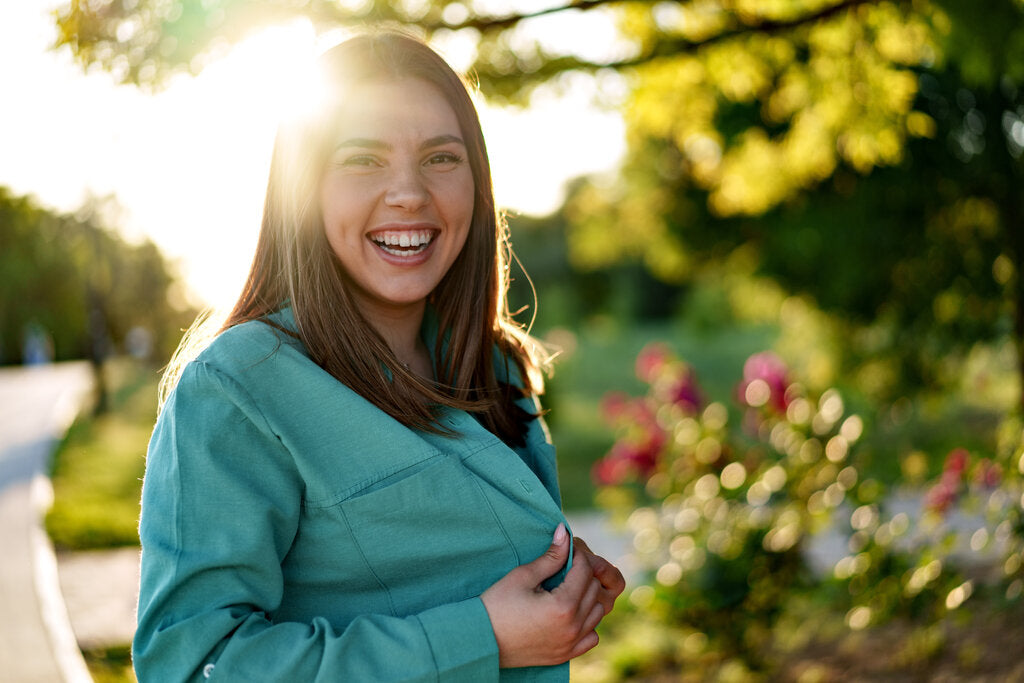 Smiling woman outdoors in the sun wearing a long-sleeve shirt, showing how UPF clothing supports skin health and sun safety.