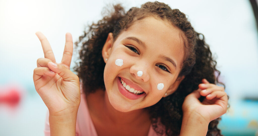 Smiling young girl with sunscreen dots on her face, flashing a peace sign. Promotes early sun safety habits with sunscreen and UPF clothing.