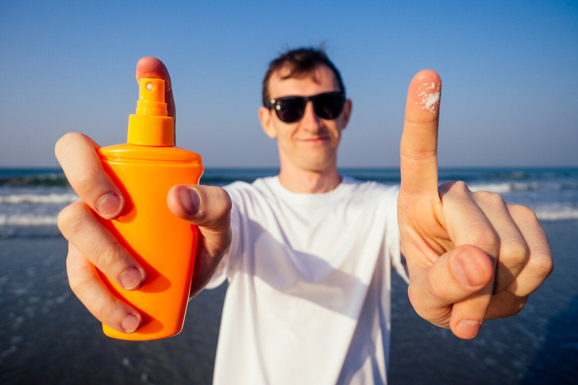 Man at the beach in a UPF t-shirt, holding a bright orange sunscreen bottle, and showing a finger with sunscreen applied, wearing sunglasses and smiling under the sun.