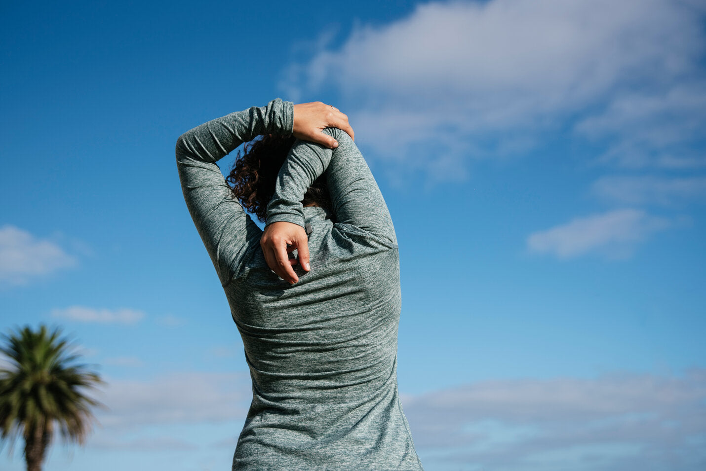 Back view of a woman stretching her arms outdoors under a clear blue sky, wearing a long-sleeve UPF-rated shirt for sun protection.