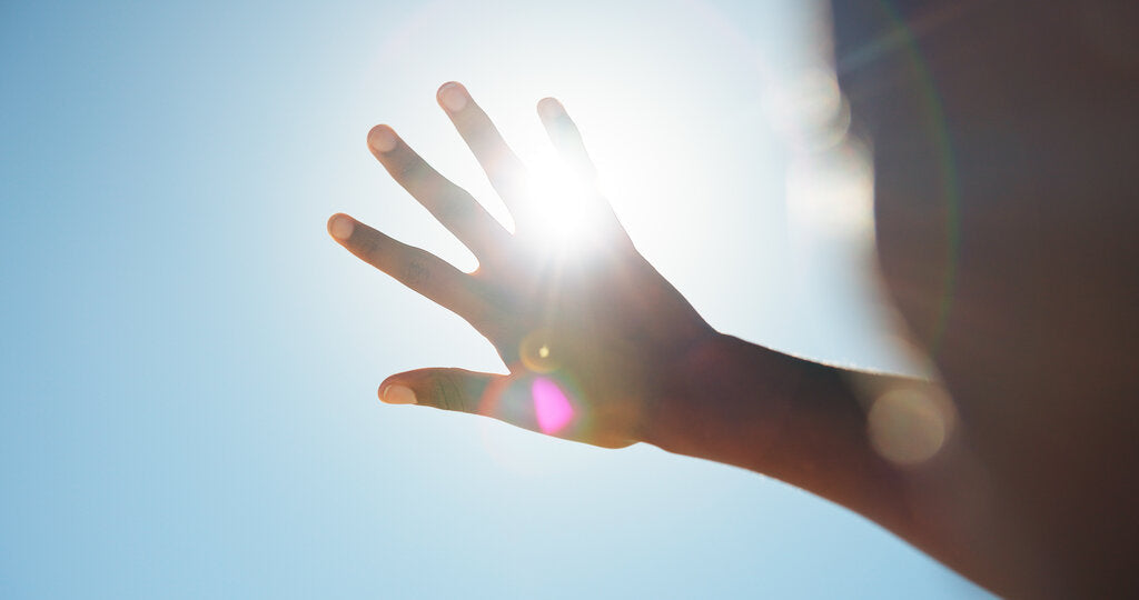 Hand raised against a bright sun in a clear blue sky, sunlight streaming between the fingers.