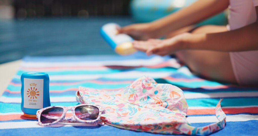 Child applying sunscreen on a beach towel next to sunglasses, a sun hat, and SPF products, showing how multiple sun protection essentials work together against UV rays