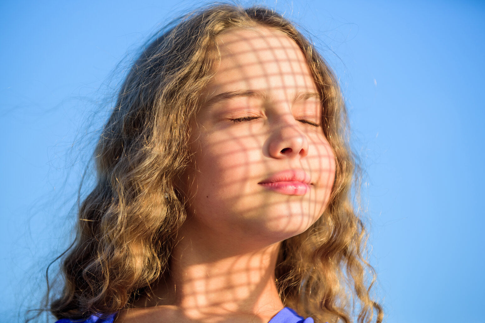 Young girl with closed eyes under a grid shadow, symbolizing the unseen effects of UVA and UVB rays and the importance of daily sun protection.
