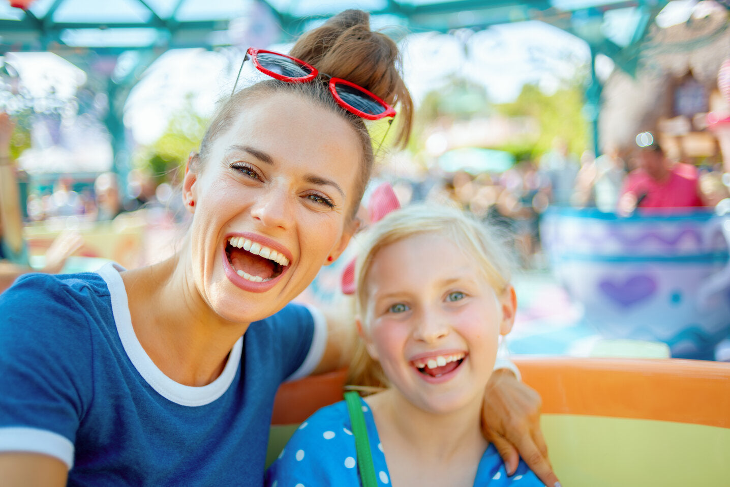 A cheerful woman and a young girl smiling and enjoying a ride at an amusement park. The woman is wearing sunglasses on her head, and both are laughing in a fun, colorful environment.