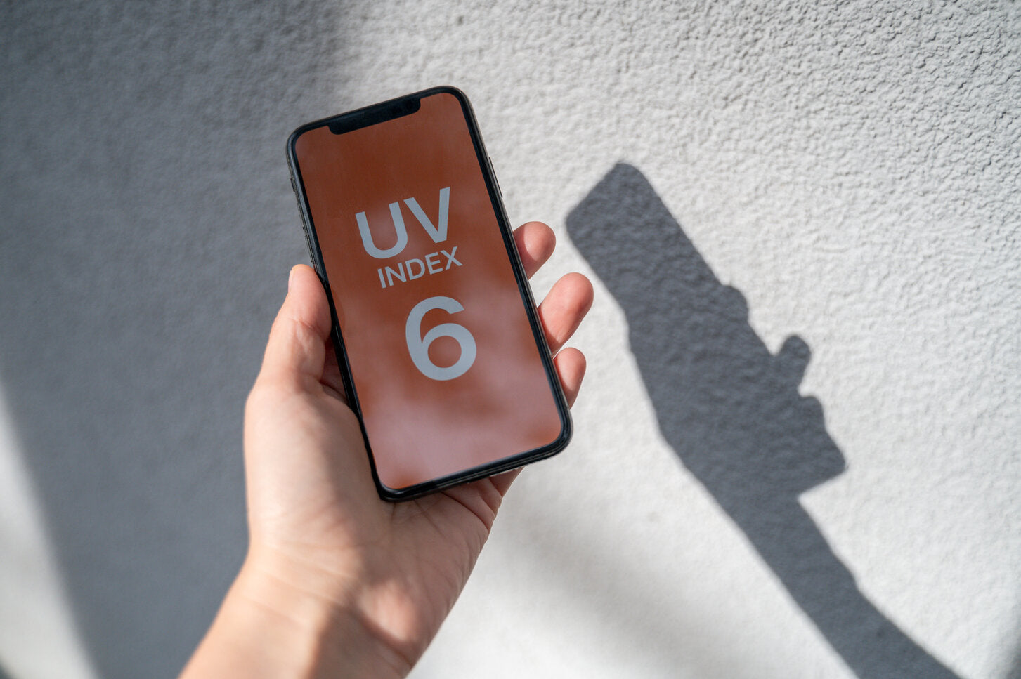 Person holding a smartphone displaying UV index 6, with sunlight casting a shadow on the wall.