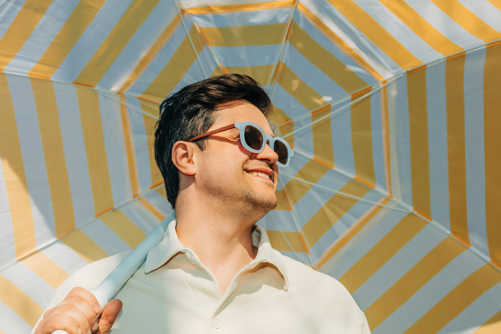 Person standing beneath a yellow and white striped beach umbrella while wearing sunglasses, illustrating how shade with ultraviolet effectiveness (UVE) reduces UV exposure.