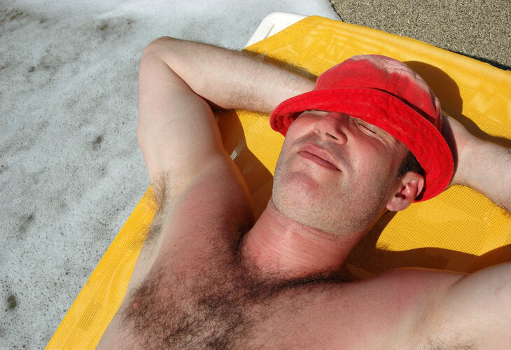 Man lying in the sun wearing a red hat, with visible sunburn on his chest and shoulders, illustrating what it means to be prone to sunburn during unprotected UV exposure.