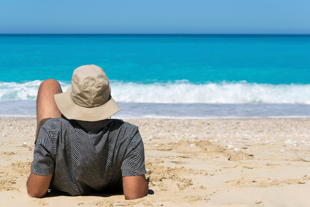 Man relaxing on the beach wearing a wide-brimmed hat and UPF 50+ shirt, showing how sun-protective clothing helps prevent sunburn and skin damage from strong UV rays.