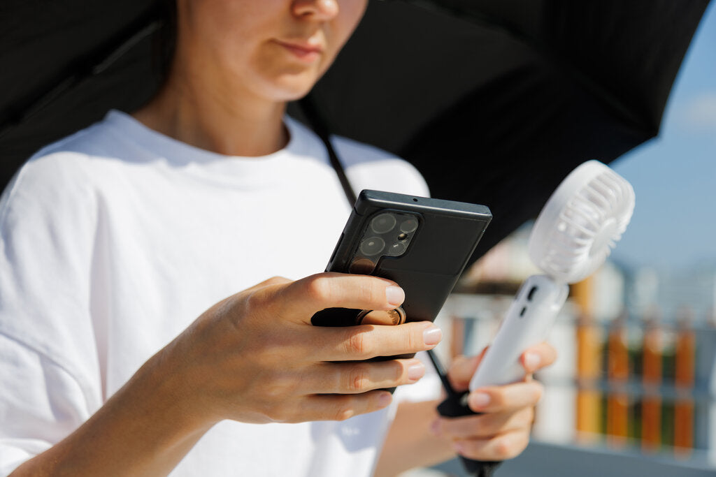 Woman shaded by an umbrella checking the UV Index on her phone while holding a portable fan during a heat wave
