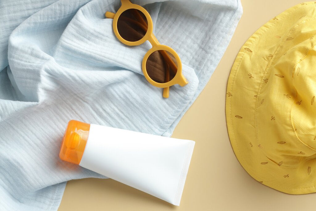 Flat lay of sun protection essentials: an unlabeled sunscreen tube, yellow-framed sunglasses, a yellow sun hat, and a light blue cloth, the everyday pieces UPF and SPF labels describe.