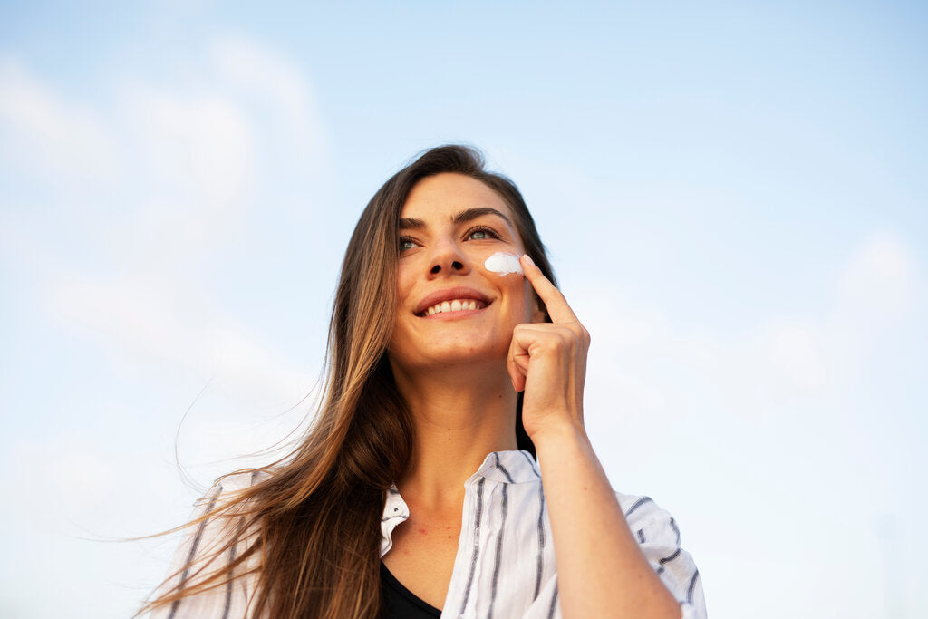 Smiling woman applying sunscreen while wearing a lightweight shirt, representing a smart sun protection wardrobe that combines skincare and protective clothing.
